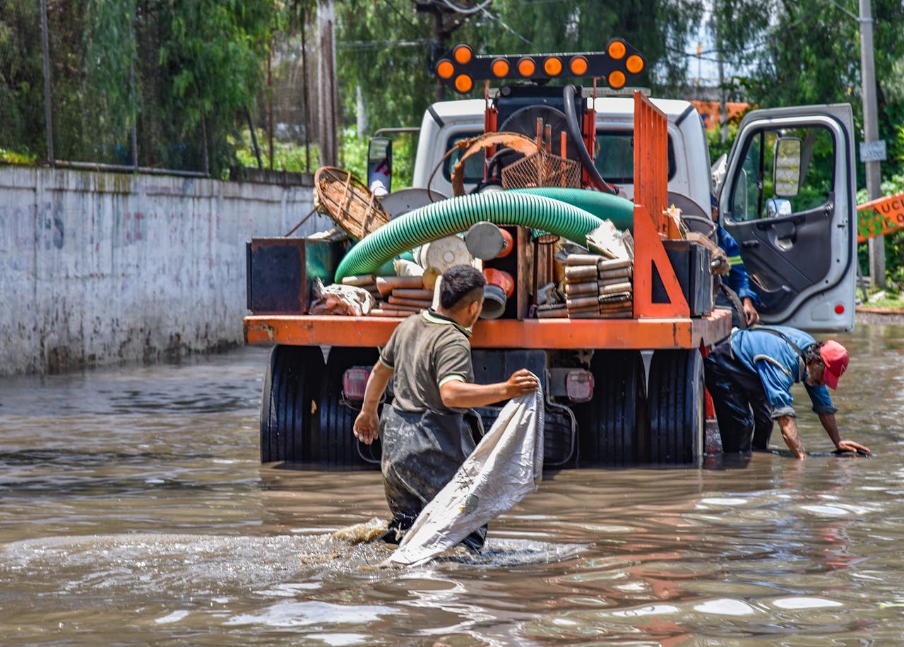 hero-img-01 Workers wading through floodwaters near a truck, managing a city crisis after heavy rain.