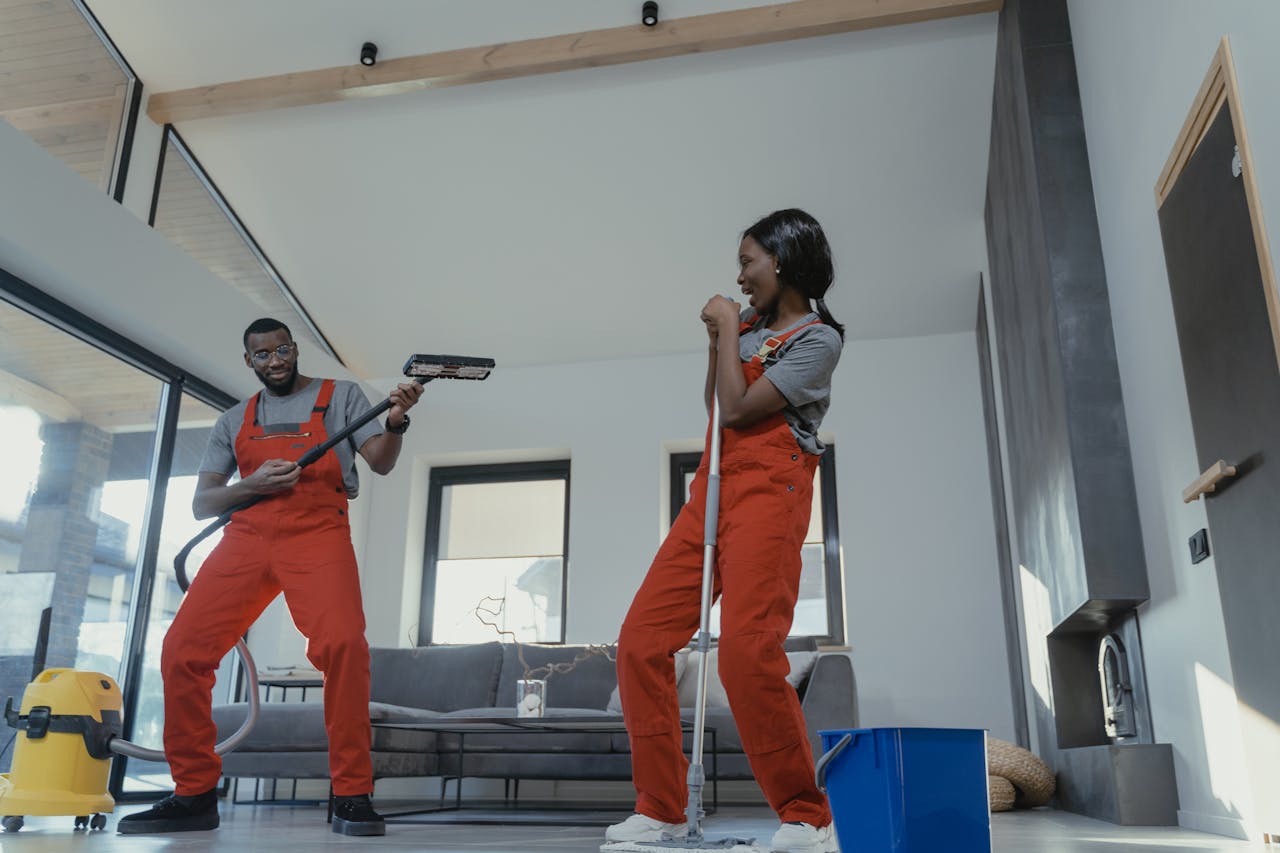 hero-img-02 Two adults in vibrant coveralls cleaning a modern home interior, enjoying work.